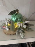 Light-up green bauble Christmas centrepiece on a wood slice with snowy greenery, white berries and a miniature tree